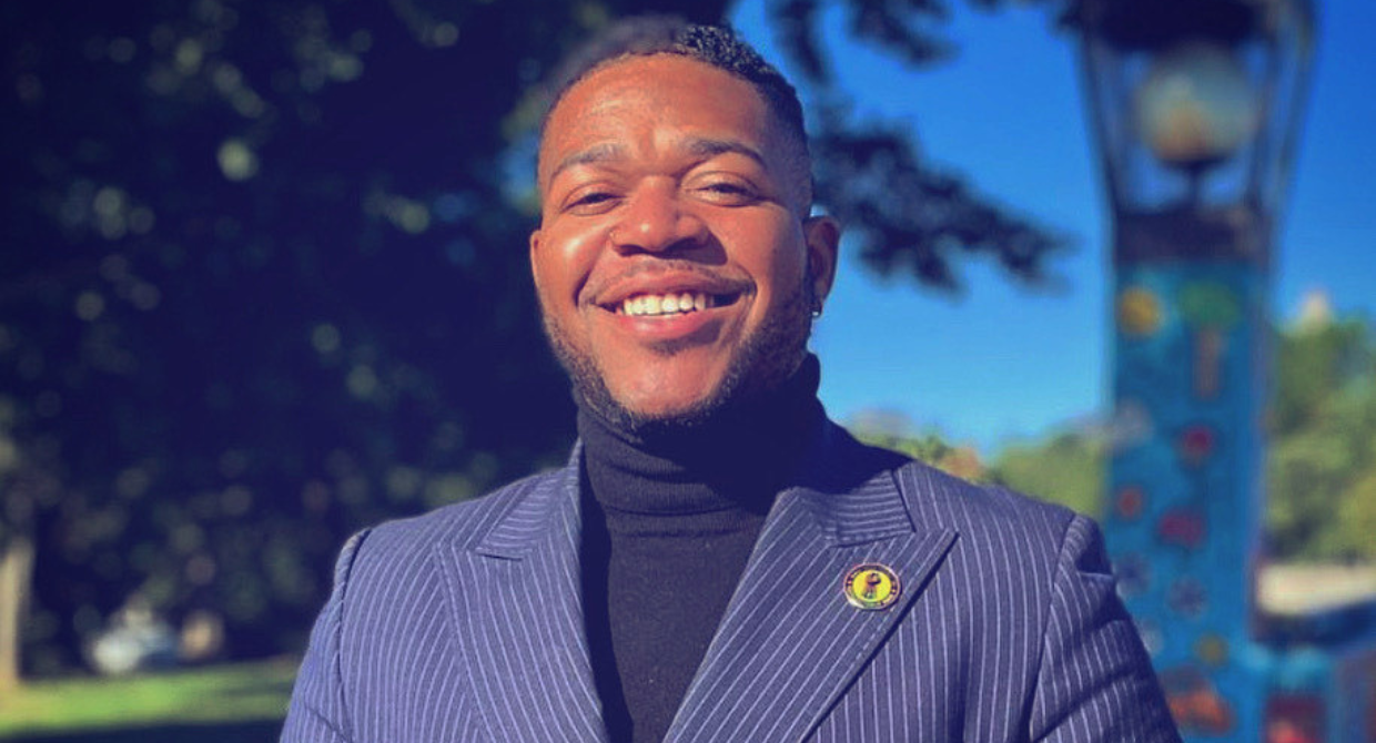 A Black person wearing a black turtleneck and blue blazer with a Black Youth Project 100 lapel pin smiles at the camera. Trees and grass are visible behind him.